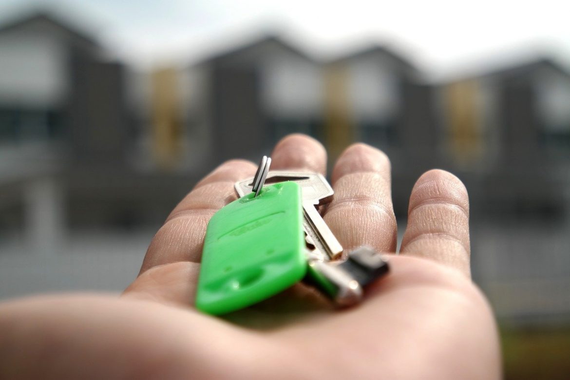 Mão aberta, com molho de chaves em um chaveiro verde, a frente imagem de casas ofuscadas