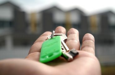 Mão aberta, com molho de chaves em um chaveiro verde, a frente imagem de casas ofuscadas